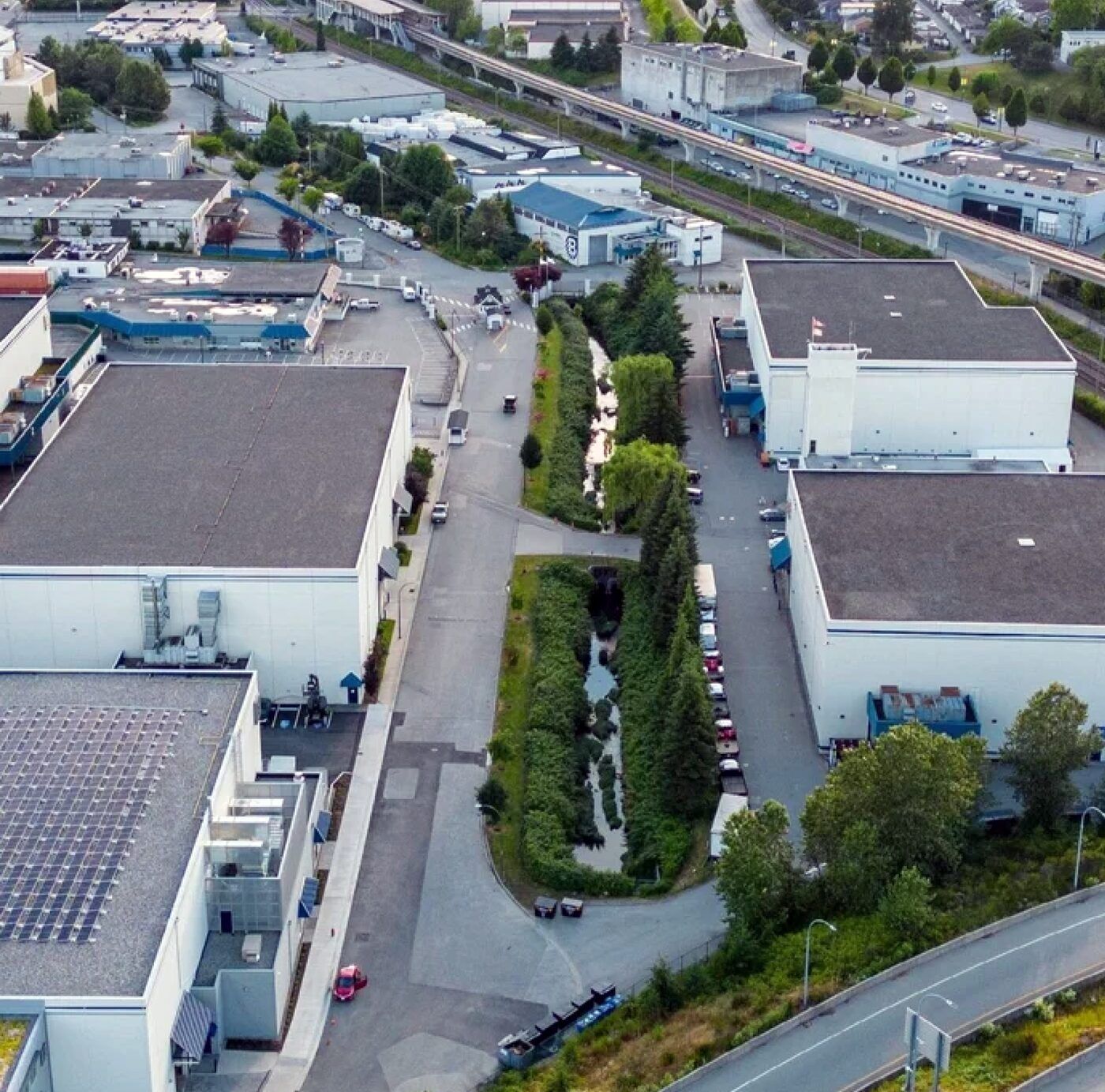 Aerial view of an urban stream running in a narrow green corridor between industrial buildings, roads, and rail lines